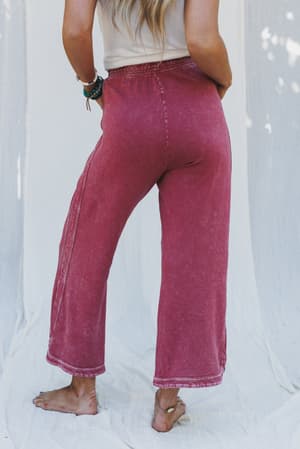 A woman stands barefoot on a white fabric backdrop, wearing the So Comfy Wide Leg Pant Cropped Length in Burgundy. The mineral-washed sweatpants have a wide leg and high-rise elastic waist. Her long hair is down, bracelets on her wrist, photographed from behind.