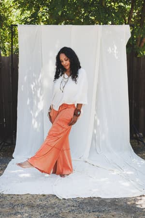 A woman with curly dark hair stands barefoot outdoors on a white cloth, wearing an oversized white blouse and the Game On Cargo Wide Leg Pant in Burnt Orange, styled with layered necklaces and bracelets. A white fabric backdrop and trees are behind her.