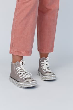 A person wears light pink cropped pants and the Chantel Studded High Top Sneaker in mocha with white laces, chunky soles, and subtle rhinestone details, standing against a plain light gray background.