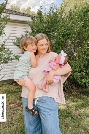 A smiling woman wears the So Comfy Wide Leg Denim Pant - Denim, featuring a flowy design and elastic drawstring waist, as she stands outdoors holding a young child and an infant. Lush greenery and a white shed create a charming backdrop.
