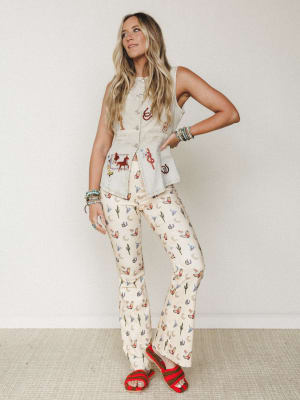 A sun-kissed blonde model poses confidently in a playful western-print flare pant and embroidered denim vest combo, styled with red crochet sandals and stacked turquoise bracelets — the kind of outfit that makes a country concert feel like a whole pe
