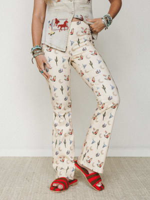 A cropped mid-shot of wide-leg flare pants in cream with an all-over western novelty print — cowboy boots, cacti, horseshoes, crescent moons, and longhorn skulls — styled with a matching embroidered denim vest and red crochet sandals, stacked turquoi