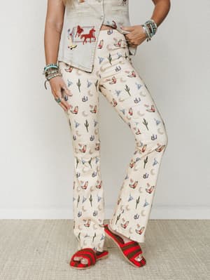 A cropped mid-shot of wide-leg flare pants in cream with an all-over western novelty print — cowboy boots, cacti, horseshoes, crescent moons, and longhorn skulls — styled with a matching embroidered denim vest and red crochet sandals, stacked turquoi