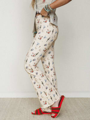 A side-profile shot of wide-leg flare pants in a cream base covered in a playful western novelty print — cowboy boots, cacti, horseshoes, and crescent moons — styled with a sage button-down, stacked turquoise bracelets, and red crochet sandals for an