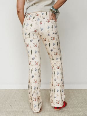 Back view of cream flare pants covered in a playful western novelty print — cowboy boots, cacti, crescent moons, horseshoes, and longhorn skulls — styled with a sage tee, stacked turquoise bracelets, and red embroidered shoes.