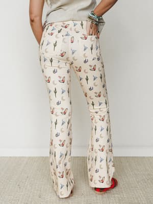 Back view of cream flare pants covered in a playful western novelty print — cowboy boots, cacti, crescent moons, horseshoes, and longhorn skulls — styled with a sage tee, stacked turquoise bracelets, and red embroidered shoes.