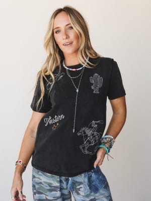 A washed black graphic tee covered in western-themed embroidery — cactus, rodeo rider, lasso, script lettering — styled with camo pants and stacked jewelry for a lived-in, free-spirited vibe.
