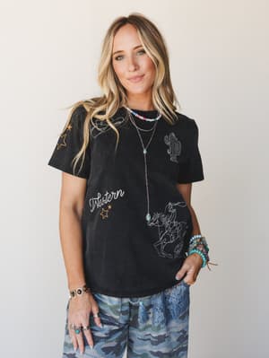 A washed black graphic tee covered in western-themed embroidery — cactus, cowboy on horseback, lasso, stars, and a cursive 'Western' script — styled with blue printed wide-leg pants and layered turquoise and beaded jewelry for a lived-in, free-spirit