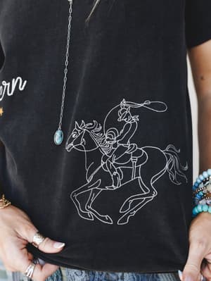 A close-up of a washed black graphic tee featuring a detailed line-art embroidery of a cowgirl on a rearing horse with a lasso, styled with turquoise jewelry, stacked bracelets, and silver rings — western energy done right.