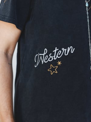 A close-up detail shot of a washed black graphic tee with 'Western' embroidered in a cursive script, accented by gold star embroidery — the kind of shirt that feels broken-in before you even buy it.