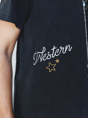 A close-up detail shot of a washed black graphic tee with 'Western' embroidered in a cursive script, accented by gold star embroidery — the kind of shirt that feels broken-in before you even buy it.