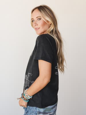 A model shot from the side showing a washed black graphic tee with a line-art cowgirl and lasso illustration, styled with stacked beaded bracelets and a peek of blue lace-trim bottoms — effortless western-boho energy.