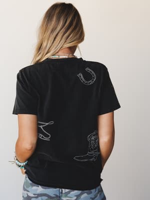 A back-view shot of a relaxed black tee with white embroidered western motifs — horseshoe, cowboy boot, and lasso — styled with camo shorts, layered bead necklaces, and a turquoise bracelet stack for that effortless western-boho vibe.