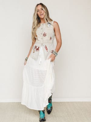 A breezy, story-filled outfit pairing a washed sand embroidered vest — dotted with western motifs like horses, lassoes, and horseshoes — over a flowy white maxi skirt, finished with statement turquoise cowboy boots and stacked bead bracelets.