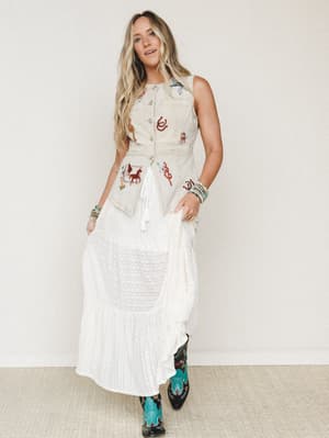 A breezy, story-filled outfit pairing a washed sand embroidered vest — dotted with western motifs like horses, lassoes, and horseshoes — over a flowy white maxi skirt, finished with statement turquoise cowboy boots and stacked bead bracelets.