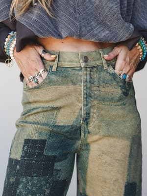 A close-up waist shot of acid-washed barrel jeans in a sandy olive and deep indigo patchwork wash, styled with a cropped gray top and layered with silver rings and turquoise beaded bracelets — the kind of jeans that look like they've been somewhere i