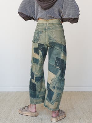 A back-view shot of wide-leg barrel jeans in a heavily distressed vintage wash, covered in patchwork denim panels with visible stitching — styled with a cropped gray hoodie and platform espadrilles for an effortlessly lived-in look.