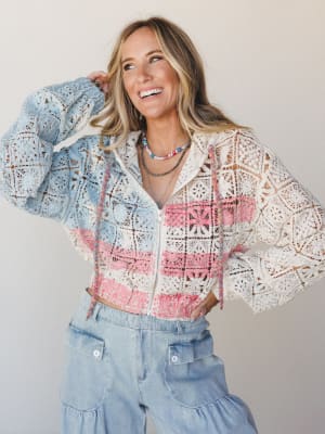 A smiling model in a cropped crochet zip-up jacket in cream, sky blue, and pink colorblock with multicolor drawstring trim, paired with light wash wide-leg denim — the kind of outfit that looks equally at home at a weekend market or a summer concert.