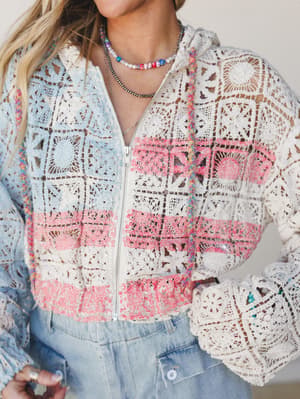 A close-up of a patchwork crochet zip-up hoodie in cream, soft pink, and sky blue granny squares, styled over light wash denim with layered beaded and pearl necklaces — the kind of piece that looks like it took someone's grandmother three summers to