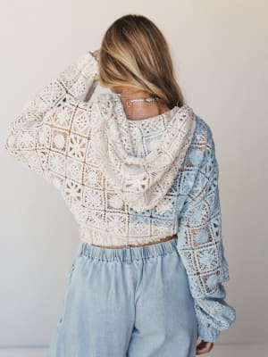 Shot from behind, a model wears a cropped crochet hoodie jacket in a two-tone colorblock of cream and dusty blue, paired with light wash wide-leg pants — the kind of effortless, textural outfit that looks like it came together on a good day.