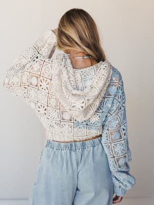 Shot from behind, a model wears a cropped crochet hoodie jacket in a two-tone colorblock of cream and dusty blue, paired with light wash wide-leg pants — the kind of effortless, textural outfit that looks like it came together on a good day.
