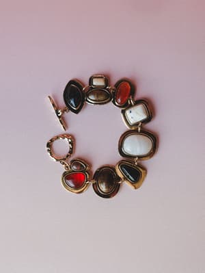 A chunky mixed-stone bracelet laid flat against a soft mauve background — each mismatched cab stone in cream, rust red, smoky brown, and black feels like something you'd find at a market and never put down.