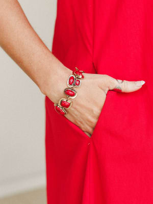 A close-up of a bold red stone and gold link bracelet worn against a red dress, hand tucked into a pocket — the kind of accessory that quietly does all the talking.