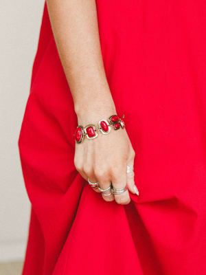 A close-up of a model's hand gathering the skirt of a bold red dress, styled with a gold link bracelet set with chunky red stones and a stack of silver rings — the kind of jewelry moment that makes an outfit feel intentional.