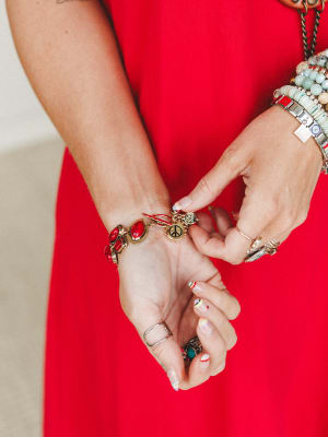 A close-up of stacked wrists dripping in personality — the red stone charm bracelet takes center stage against a bold red dress, surrounded by beaded bracelets, silver rings, and an evil eye charm that all feel collected, not coordinated.