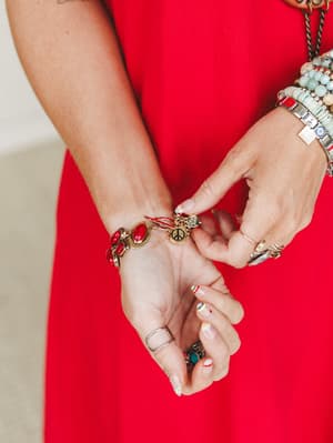 A close-up of stacked wrists dripping in personality — the red stone charm bracelet takes center stage against a bold red dress, surrounded by beaded bracelets, silver rings, and an evil eye charm that all feel collected, not coordinated.