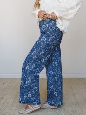 A side-angle shot of wide-leg denim jeans in a medium wash with an all-over western-inspired toile print — horses, cacti, and line-drawn motifs — styled with a cream eyelet lace top, stacked silver and turquoise rings, and woven flats.