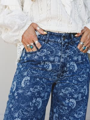 A close-up torso shot of wide-leg medium wash jeans covered in a white line-art western print — horses, cowboy boots, cacti, florals — paired with a white eyelet and lace blouse, hands tucked in pockets and stacked with silver and turquoise rings.