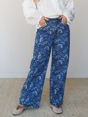 Wide-leg denim with an all-over western toile print — horses, cacti, cowboy boots rendered in soft white linework on medium indigo — styled with a ruffled lace blouse and woven flats, hands in pockets, rings stacked.