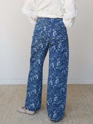 A back-view shot of wide-leg denim jeans in a medium indigo wash printed with a whimsical western toile — horses, cacti, cowboy boots — paired with a cream lace-trim blouse and woven flats for an effortlessly boho, Western-inspired look.