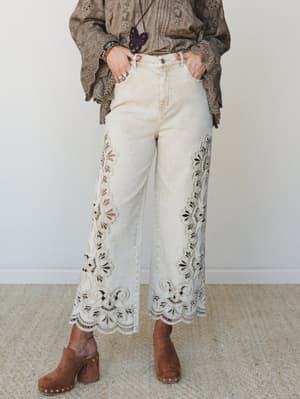 Wide-leg crop jeans in washed sand denim steal the show with sweeping eyelet cutwork and scalloped hem — styled with a taupe eyelet blouse, stacked bracelets, and suede clogs for a look that feels vintage and very much intentional.