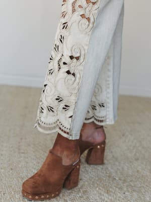 A close-up detail shot of wide-leg sand-toned jeans with intricate eyelet embroidery and scalloped hem, paired with cognac suede platform clogs on a natural jute rug — the kind of detail that makes someone stop scrolling.