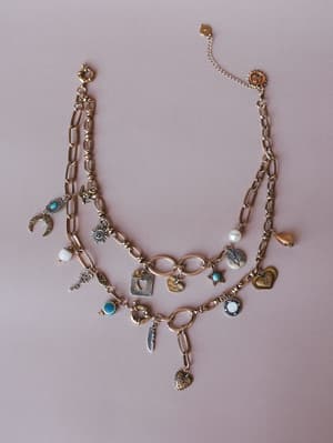 A layered gold chain charm necklace styled flat on a dusty rose background, loaded with an eclectic mix of symbolic charms — moons, hearts, a key, feather, sun, pearl, and turquoise accents — that feels like a collection of little stories worn all at