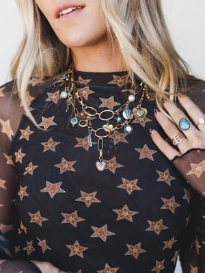 A close-up styled shot showcasing layered gold chain necklaces loaded with mixed charms — pearls, turquoise, hearts, feathers, and coins — worn over a sheer black mesh top with warm brown star print, finished with a stack of silver and turquoise ring