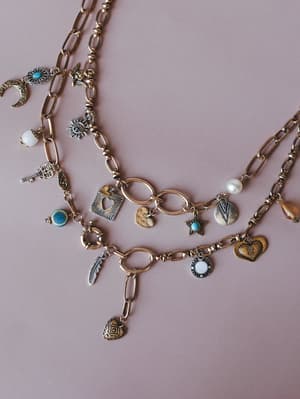 A close-up styled flat lay of layered gold chain charm necklaces arranged on a dusty rose surface, each strand loaded with mixed-metal charms — moons, hearts, feathers, keys, stars, and turquoise stones — giving off serious collected-over-time energy