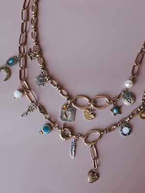 A close-up flat lay of layered gold chain necklaces loaded with mixed-metal charms — moons, suns, hearts, keys, feathers, turquoise stones, and a pearl — styled against a dusty mauve background with the kind of collected, meaningful energy of jewelry