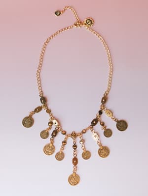 A gold coin charm necklace laid flat against a soft pink gradient background — the kind of piece that looks like it traveled somewhere interesting and has stories to tell.
