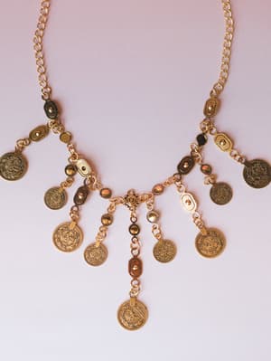 A gold coin charm necklace laid flat against a soft blush background — the kind of piece that looks like it has a story, with engraved medallion pendants cascading in a draped bib silhouette.