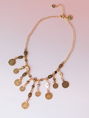 A gold coin charm necklace laid flat against a soft pink gradient background — the kind of piece that looks like it traveled somewhere interesting and has stories to tell.