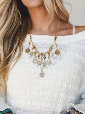 A close-up lifestyle shot of a layered gold coin charm necklace worn over a white open-knit sweater — boho and easy, the kind of piece that makes a simple outfit feel like it has a story.