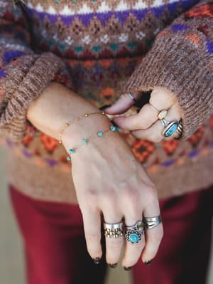 A close-up of stacked hands layered in silver and turquoise rings alongside a delicate gold chain bracelet with turquoise stone drops, all set against the cozy texture of a fair isle sweater — the kind of jewelry moment that looks collected, not coor