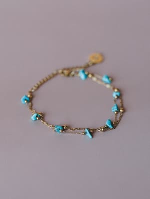 A delicate double-strand gold chain bracelet scattered with raw turquoise stone chips and tiny gold beads, shot flat against a soft lavender backdrop — the kind of piece that looks like it never comes off.