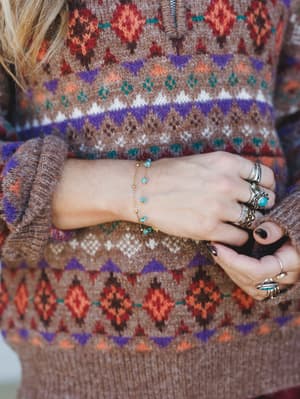 A close-up of stacked silver rings and a delicate turquoise-stone gold chain bracelet layered against a hand resting on a cozy fair isle knit sweater — the kind of jewelry moment that feels collected over time, not purchased all at once.