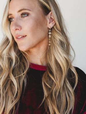 A close-up portrait shot showcasing long gold drop earrings with mixed gemstone details against a model in a deep burgundy velvet top — the whole thing has a warm, collected holiday-party energy.