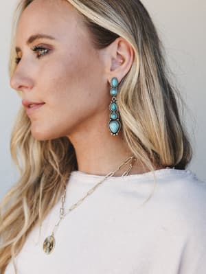 A close profile shot of a model wearing statement turquoise and silver drop earrings layered with a gold paperclip chain and coin pendant necklace — the kind of jewelry stack that looks effortless but definitely wasn't an accident.