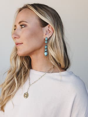 A three-quarter profile shot of a model wearing turquoise and silver drop earrings layered with a gold coin pendant necklace — the kind of jewelry combination that looks effortless but is definitely intentional.